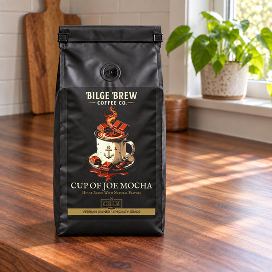 A black coffee bag labeled 'Bilge Brew Coffee Co. Cup of Joe Mocha' stands on a wooden table in a bright kitchen with a window, plant, and cutting board in the background.