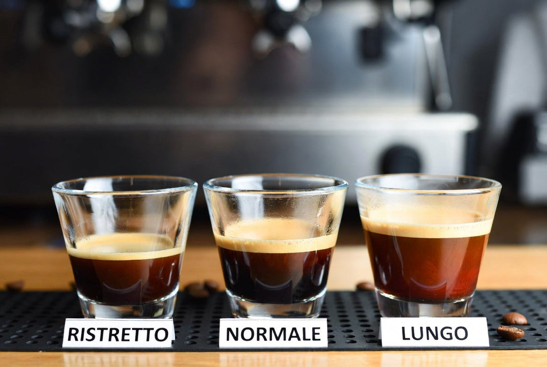 A photograph of three espresso shots (ristretto, normale, lungo) in clear cups, comparing crema and volume in a blurred coffee bar setting.