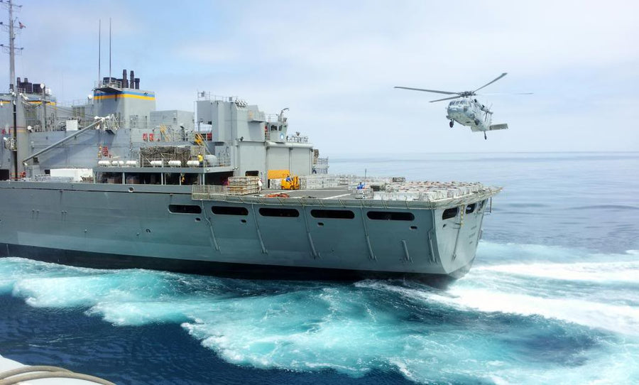 A large gray military ship sailing in open ocean with a helicopter flying overhead in the sky.