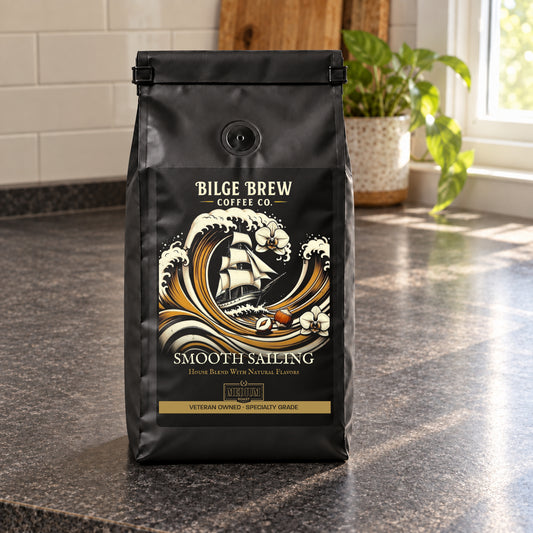 A black bag of Bilge Brew Coffee Co. 'Smooth Sailing' house blend coffee sits on a dark granite countertop in a kitchen setting with a tiled backsplash and a potted plant in the background.