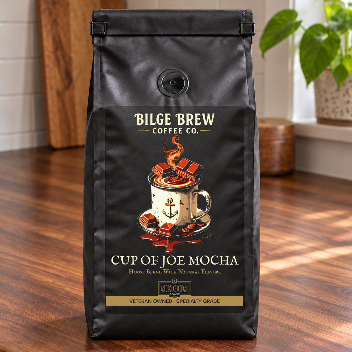 A black coffee bag labeled 'Bilge Brew Coffee Co. Cup of Joe Mocha' stands on a wooden table in a bright kitchen with a window, plant, and cutting board in the background.