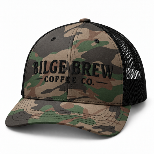 A camouflage trucker hat with black mesh back panels and black embroidered text reading 'BILGE BREW COFFEE CO.' on the front, shown on a clean white background.