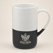 A ceramic coffee mug with a two-tone design, featuring a white upper half with subtle black speckles and a black lower half. The mug is printed with a white anchor logo and the text 'BILGE BREW COFFEE CO.