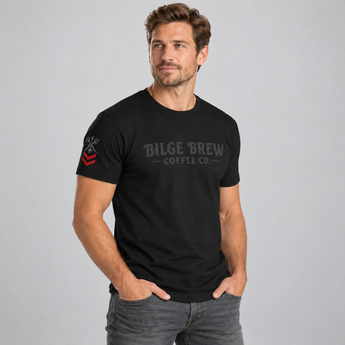 A man wearing a black crewneck t-shirt with a gray 'Bilge Brew Coffee Co.' graphic on the chest and a red and black sleeve graphic, posed against a solid gray studio background.