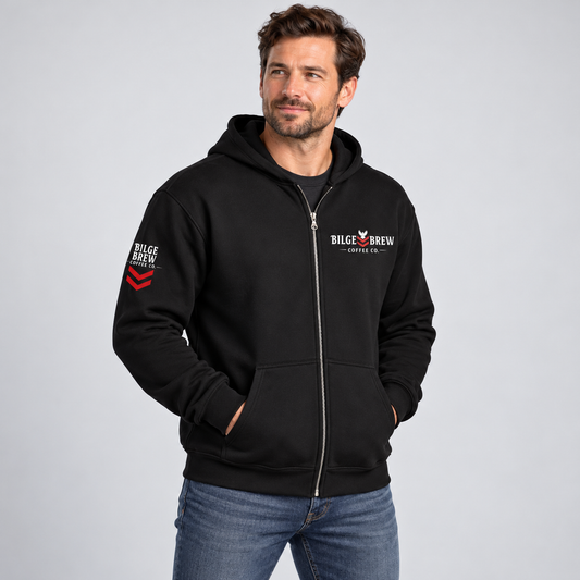 A man wearing a black zip-up hoodie with 'BILGE BREW COFFEE CO.' printed on the left chest and right sleeve, paired with blue jeans, against a light gray studio background.