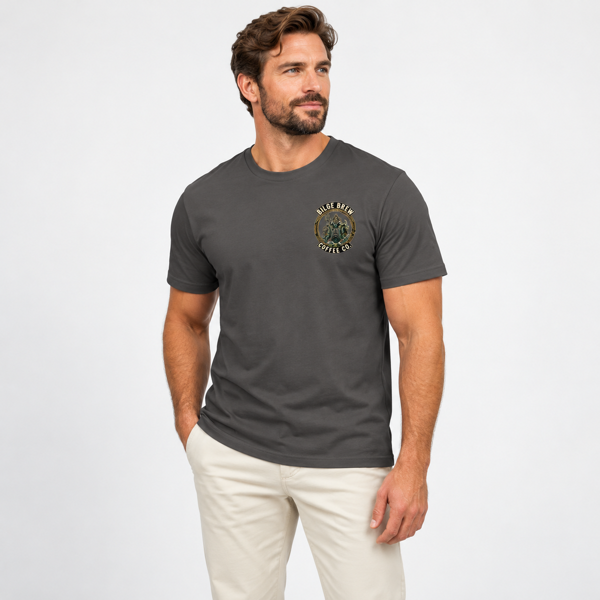 A man wearing a dark gray crewneck t-shirt with a circular 'Bilge Brew Coffee Co' graphic on the left chest, paired with light beige pants, photographed against a clean white studio background.