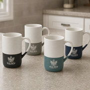Four ceramic coffee mugs with a two-tone design are arranged on a kitchen countertop. Each mug has a white upper half and a contrasting lower half in black, gray, teal, or navy, featuring a white printed anchor logo and the text 'BILGE BREW COFFEE CO.
