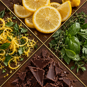 A top-down styled arrangement of lemon slices, lemon zest, fresh green herbs, and broken dark chocolate on a dark wooden surface divided by diagonal gold lines.