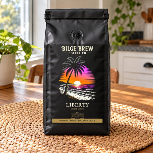 A black coffee bag standing upright on a woven placemat on a wooden table, with a bright kitchen and plants blurred in the background. The bag reads Bilge Brew Coffee Co.