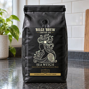 A black coffee bag labeled 'Bilge Brew Coffee Co. Sea Witch' stands on a dark countertop in a kitchen setting with a tiled wall and a plant in the background.