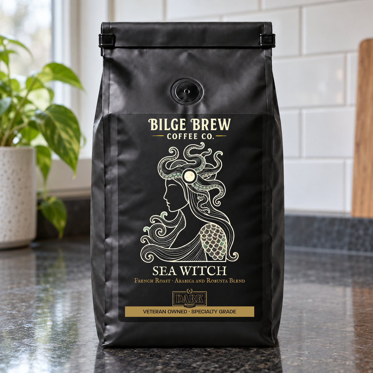 A black coffee bag labeled 'Bilge Brew Coffee Co. Sea Witch' stands on a dark countertop in a kitchen setting with a tiled wall and a plant in the background.