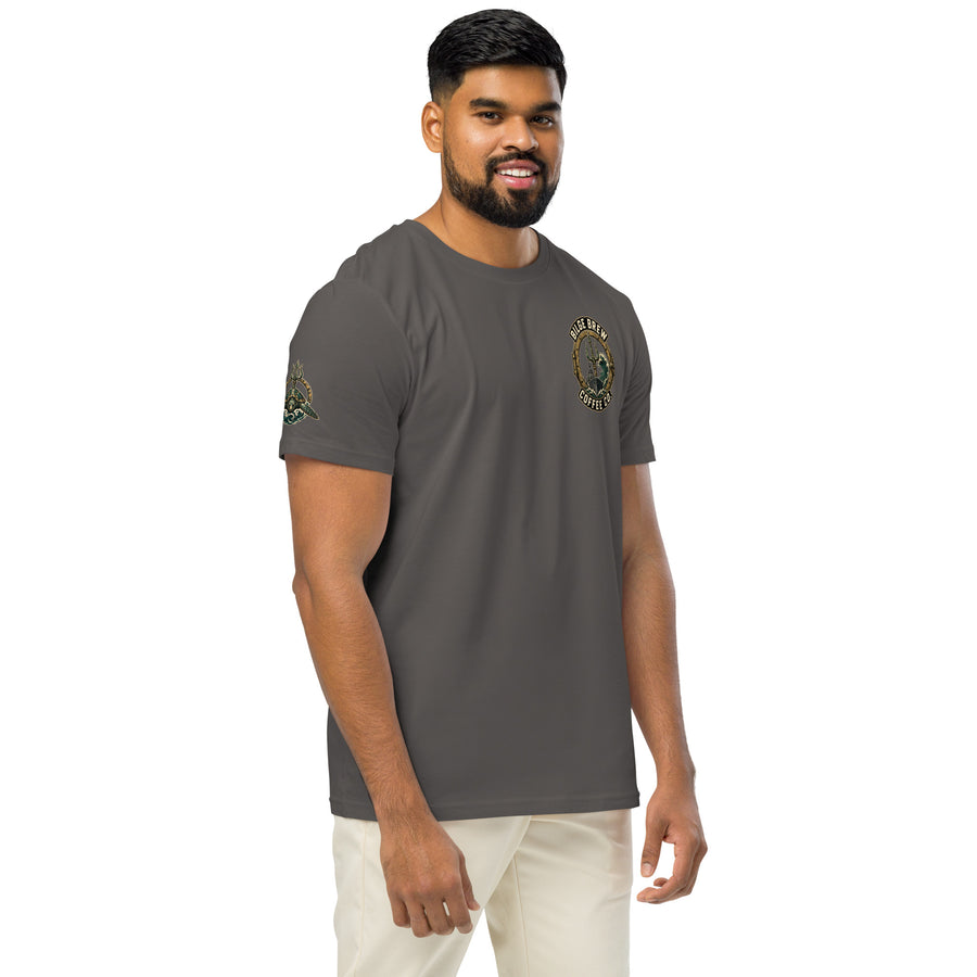 A man wearing a dark gray crewneck t-shirt with a circular military-style emblem on the left chest and a smaller emblem on the right sleeve, paired with off-white pants, set against a plain white background.